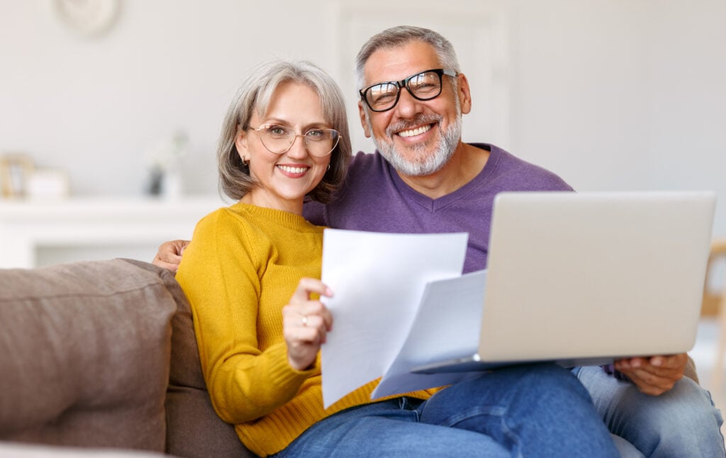 A smiling couple sits on a couch with a laptop and papers. The woman wears a yellow sweater, and the man is in a purple sweater. They both have glasses.