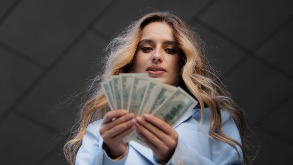 A woman with long blonde hair in a light blue blazer is holding and looking at a fan of U.S. dollar bills.