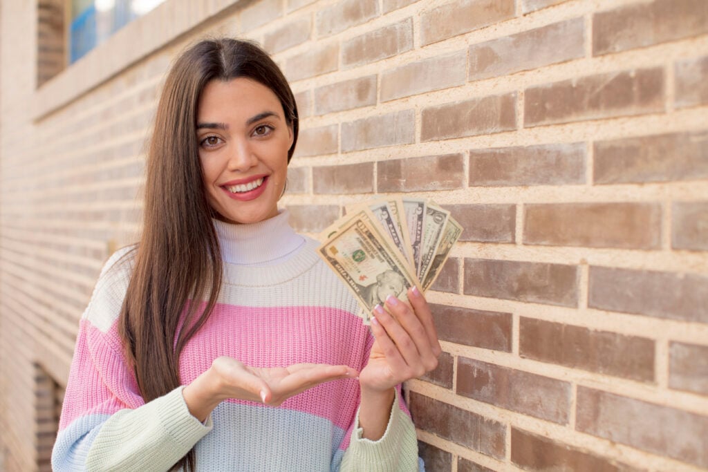 A woman with long hair smiles while holding a fan of U.S. dollar bills against a brick wall.