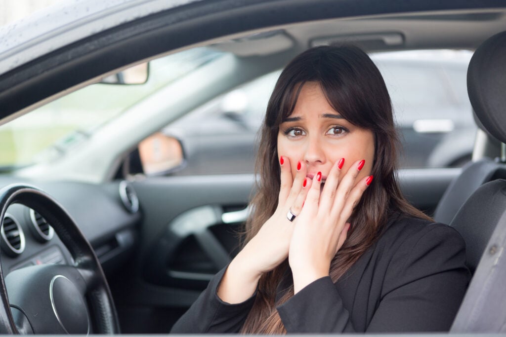 A woman with long dark hair and red nails covers her mouth with her hands while sitting in a car.