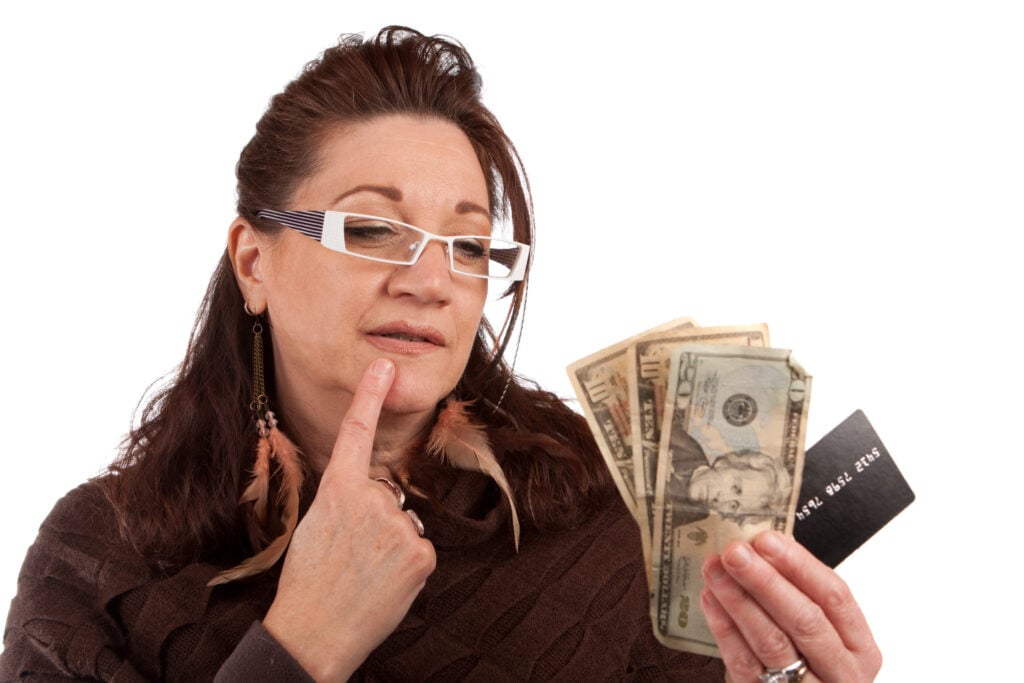 A woman wearing glasses and feather earrings is looking thoughtfully at a handful of U.S. dollar bills and a black card. She is touching her cheek with her finger.