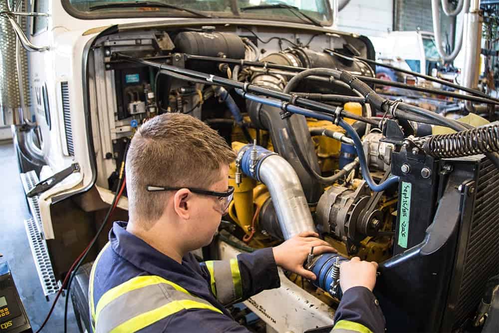 A mechanic wearing safety glasses and a uniform works on the engine of a large vehicle inside a garage.