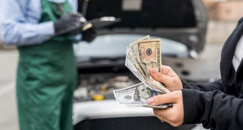 Person holding cash in front of a car with an open hood while a mechanic writes on a clipboard in the background.