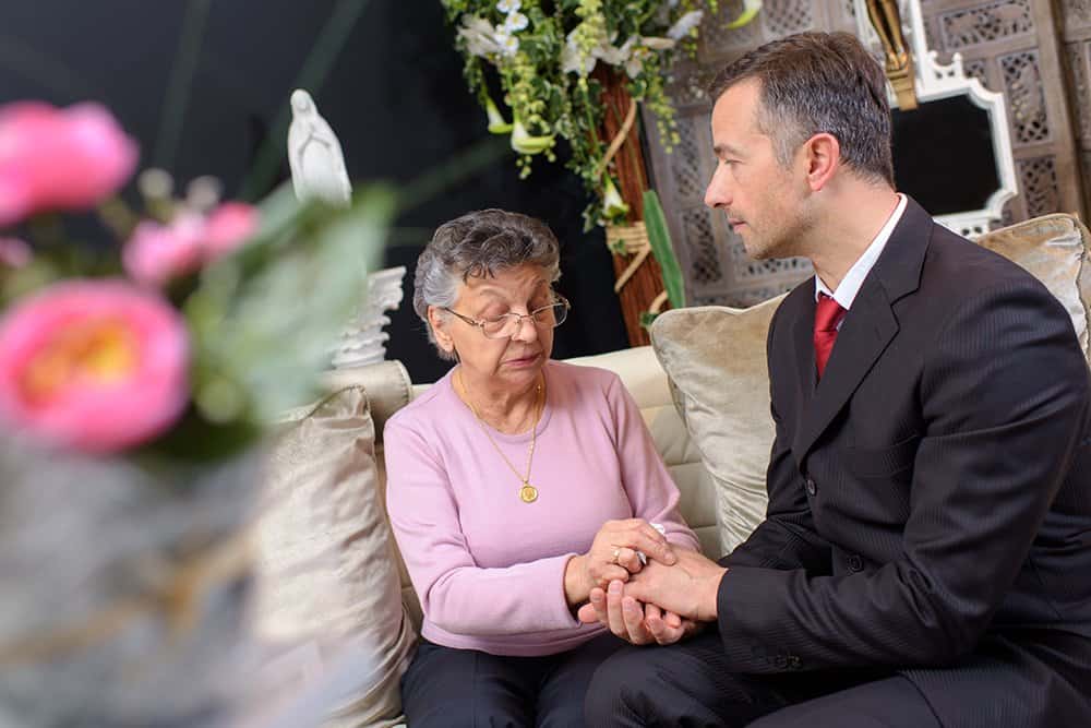 A young man in a suit holds the hand of an elderly woman wearing glasses and a pink sweater, sitting together on a couch in a room with flowers and decorative items.