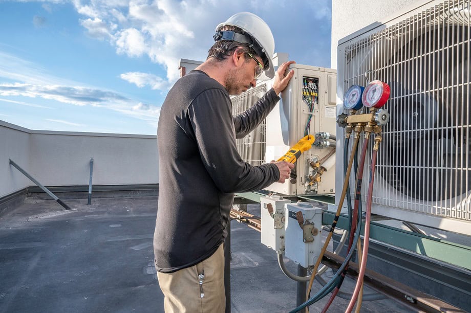 Technician in a hard hat and safety gear inspecting an air conditioning unit on a rooftop.