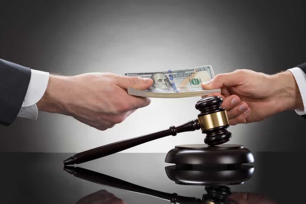 One hand passing a stack of hundred-dollar bills to another hand, with a gavel and block in the foreground.