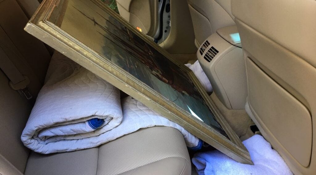 A framed painting rests on blankets in the back seat of a car.