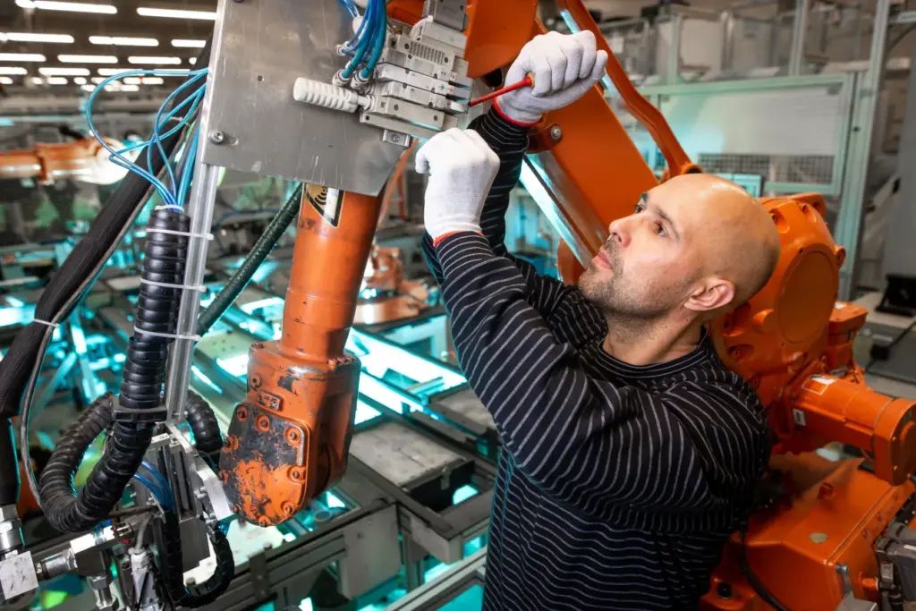 A man in a striped shirt and gloves works on an orange robotic arm in a factory setting.