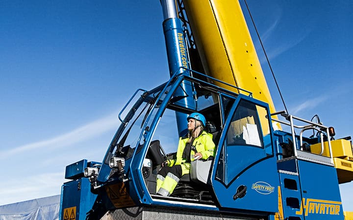 A person in a safety suit and helmet operates a blue and yellow crane under a clear blue sky.