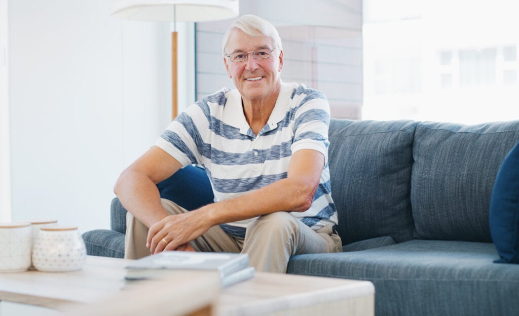 An elderly man with glasses and gray hair sits on a dark gray couch in a brightly lit room, smiling and dressed in a striped polo shirt and khaki shorts. Showcasing the independence of single seniors in America, he enjoys a moment of tranquility. A table with decor is in the foreground.