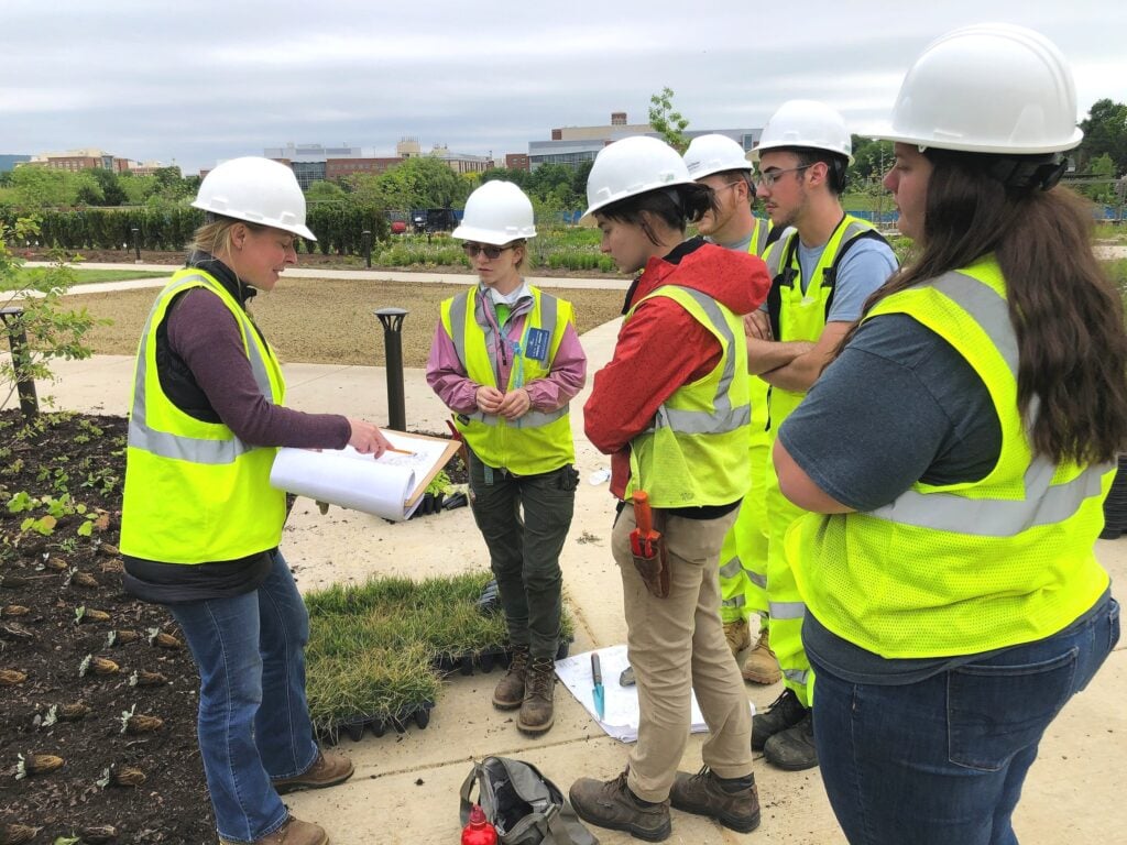 Group of people in safety vests and hard hats gathered outdoors, looking at a set of plans held by one person.