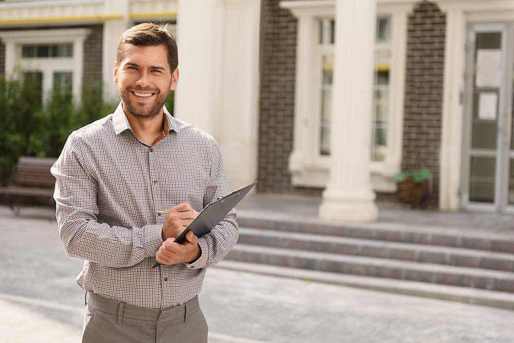 A man in a checked shirt stands outdoors holding a clipboard, smiling at the camera. In the background, there are stairs and columns leading to a brick building.