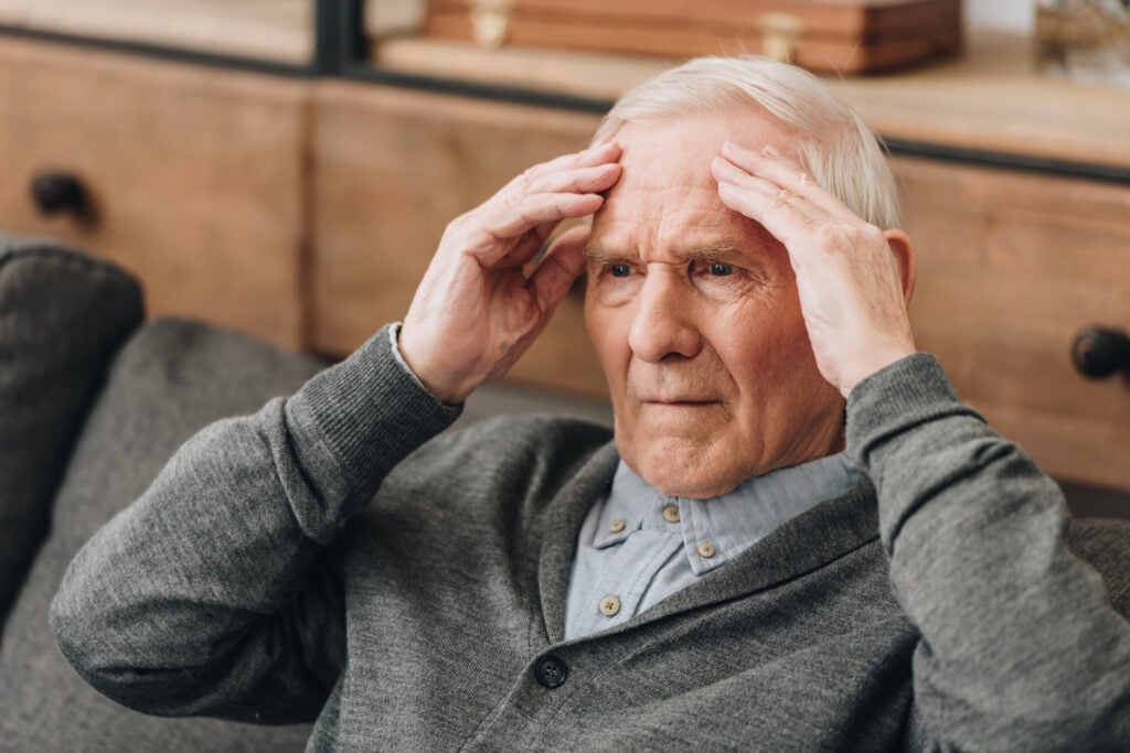 An elderly man sitting on a sofa holds his head with both hands, appearing concerned or distressed, perhaps reflecting on the advice to stop doing these if over 60.