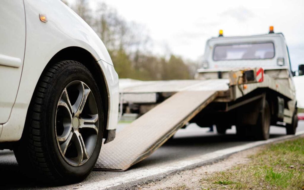 A white car is about to be loaded onto a tow truck with an extended ramp on a roadside.