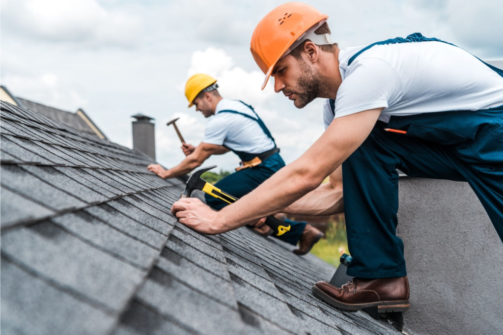 Two construction workers in helmets are installing shingles on a sloped roof.