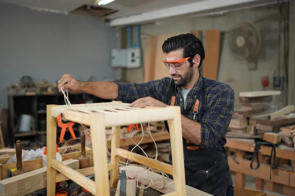 A man wearing safety glasses and an apron weaves a seat on a wooden chair in a workshop filled with tools and materials.