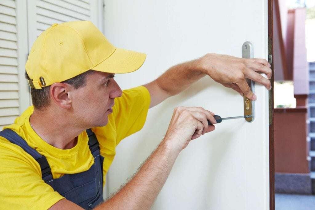 A worker in a yellow cap and shirt uses a screwdriver to fix a door lock.