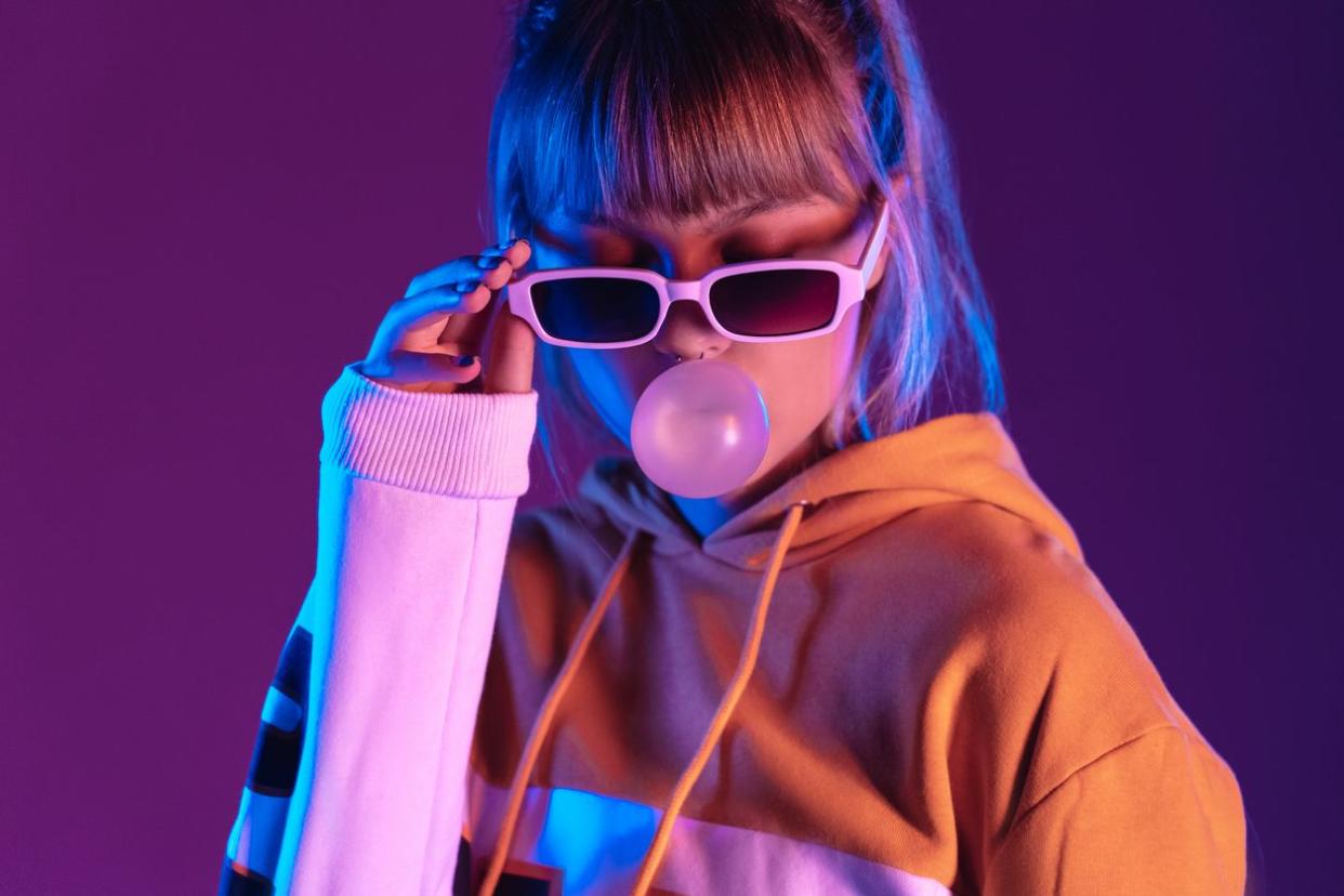 Pretty young 20s fashion teen girl model wear stylish glasses hoodie blowing bubble gum standing at purple violet studio wall background, igen teenager in trendy night party glow 80s 90s concept