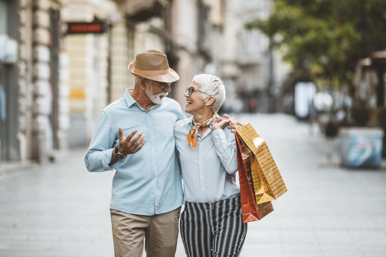 Senior Couple, Casually Dressed, is Walking in the City Center with Shopping Bags. Mature Handsome Man with Hat and Sunglasses and Grey Beard is Walking with his Beautiful Wife with Grey Hair and Talking About the Successful Shopping.