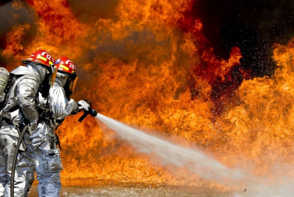 Two firefighters in protective gear are using a hose to douse large, intense flames.