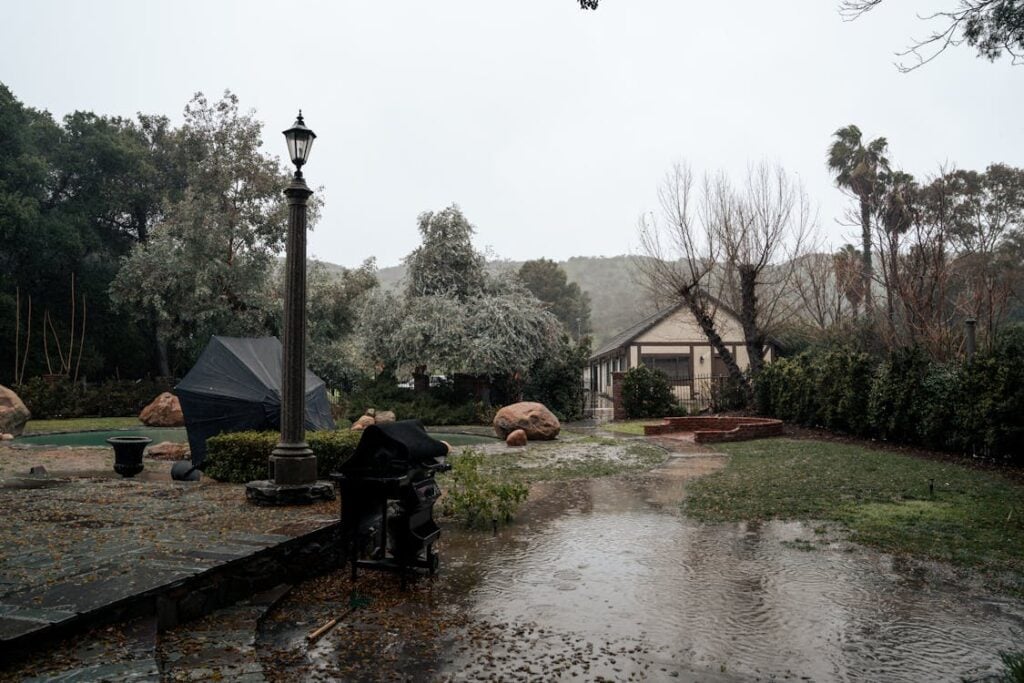 Flooded backyard with standing water, garden furniture, trees, and a small house in the background under an overcast sky.