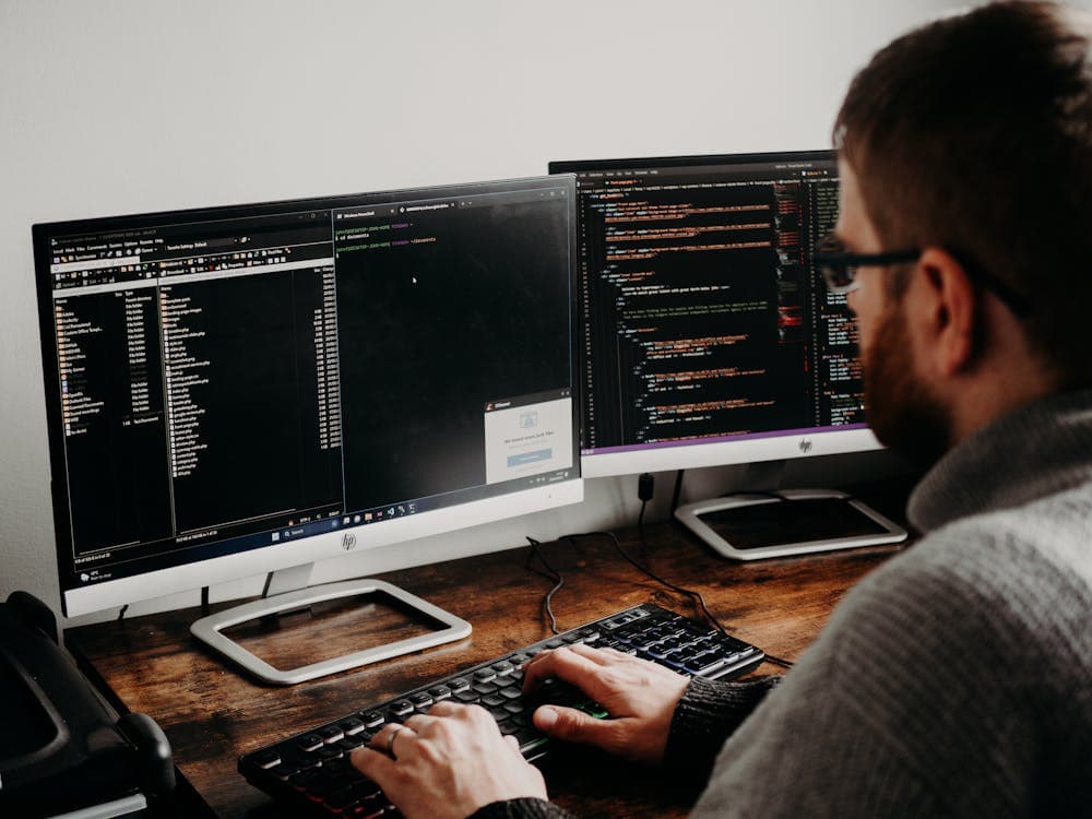 A person working on a computer with two monitors displaying code and a directory.