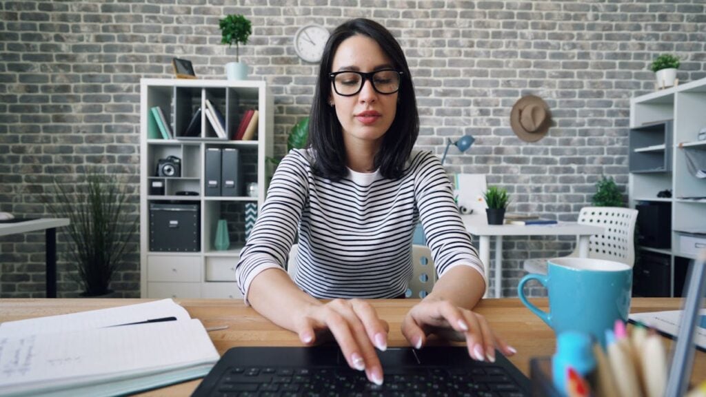 A woman wearing glasses and a striped shirt types on a laptop at a desk with a notebook, pen, and blue mug. Shelves, plants, and office supplies are in the background.