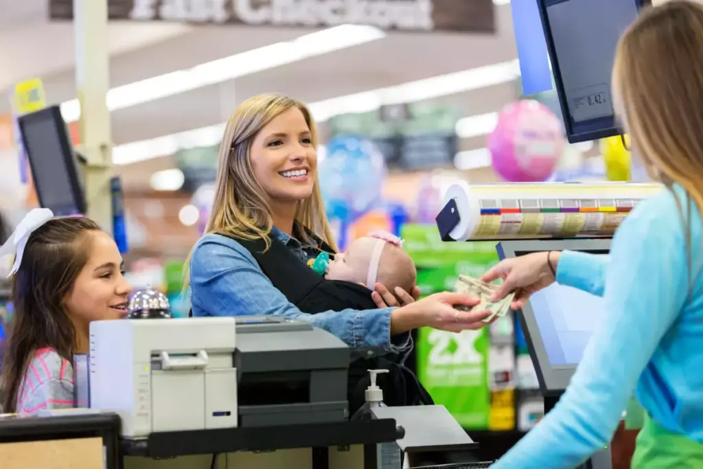 A woman holding a baby pays a cashier at a grocery store checkout, while a young girl stands nearby.