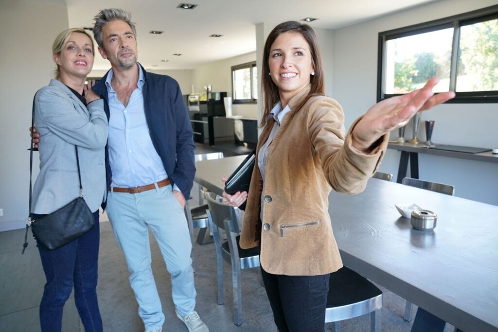 A real estate agent shows a modern home to a smiling couple. With careers in demand, the couple stands near a dining table as the agent gestures towards a part of the house, highlighting its unique features.