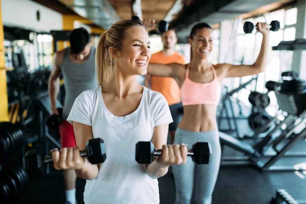 A group of people is exercising in a gym, focusing on a woman in the foreground lifting dumbbells. With others lifting weights behind her, the scene reflects the fitness lifestyle often embraced by those with an upper middle class income.