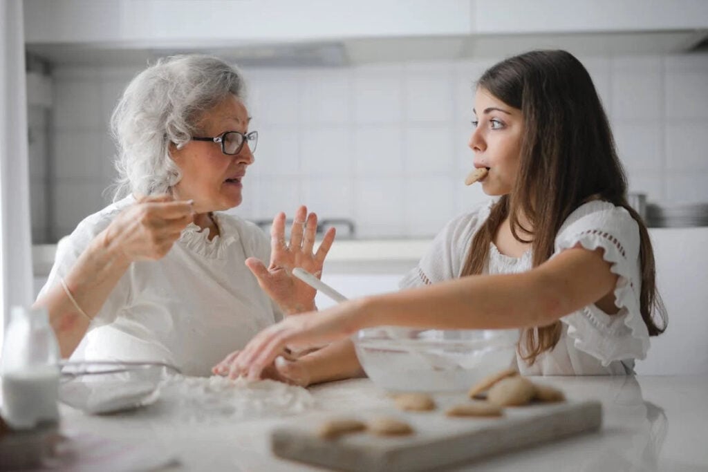 An elderly woman and a young woman are baking in a kitchen. The young woman has a cookie in her mouth while the elderly woman gestures with her hand, sharing wisdom on the hardest part of aging and how to cope. Cookies and baking ingredients are scattered on the counter.