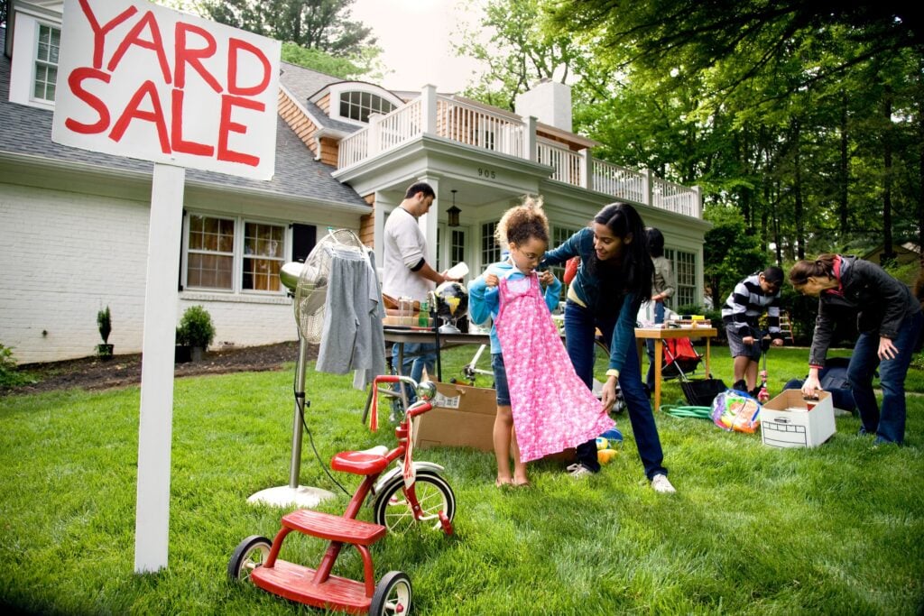 A yard sale outside a house with people browsing items. A woman helps a child try on a pink dress. A red tricycle is in the foreground.