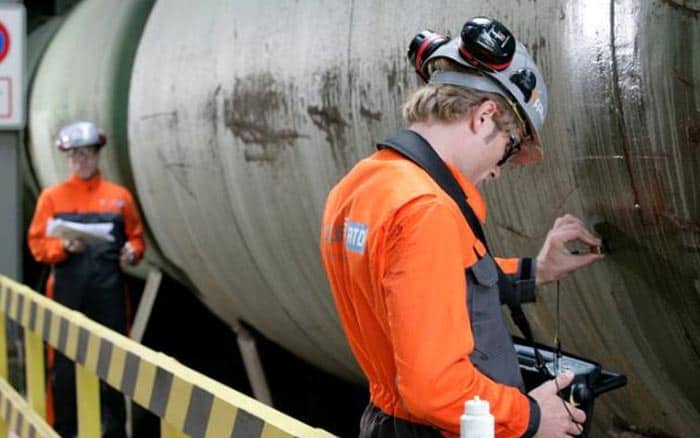 Two workers in orange uniforms and safety gear inspect a large industrial pipe, taking notes and measurements.