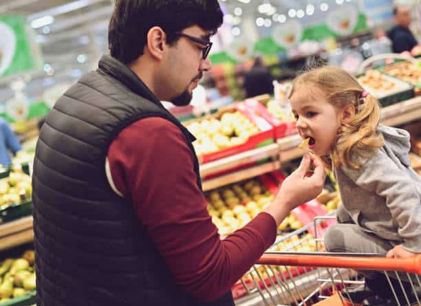 A man is feeding a small child while she sits in a shopping cart in the produce section of a grocery store.