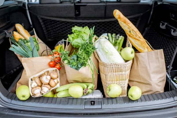 Grocery bags filled with fresh produce, including bread, leafy greens, tomatoes, mushrooms, and apples, are placed in a car trunk.