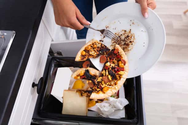 A person is scraping leftover pizza from a plate into a kitchen trash bin.