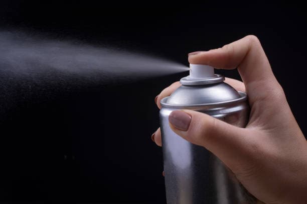 A hand holding a silver spray can, with mist spraying out against a black background.