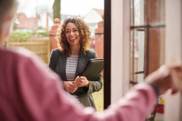 A woman with curly hair, holding a folder, stands at an open front door, smiling at an elderly person in the foreground.