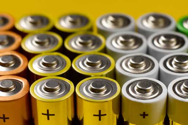 Close-up view of several rows of AA batteries aligned, with visible positive terminals. Colors of battery wrappers include yellow, white, orange, and green.