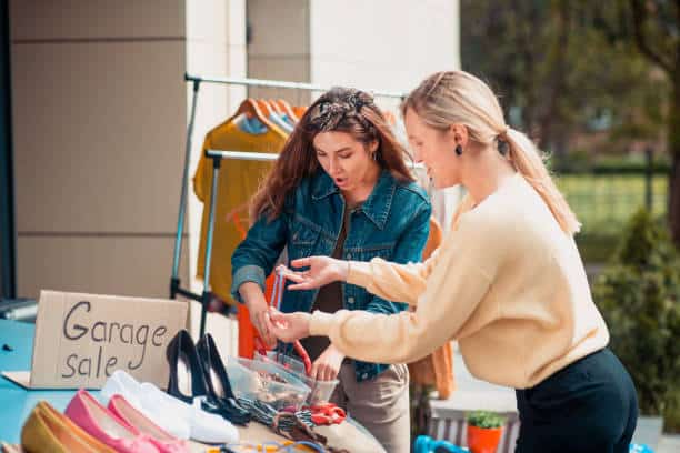 Two women at an outdoor garage sale looking through items on a table with clothes and shoes on display.