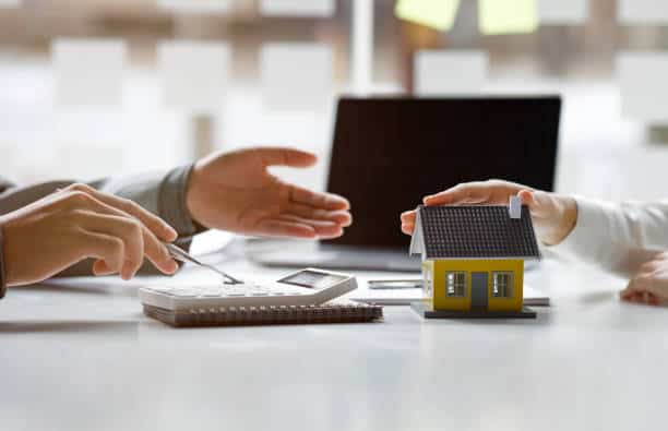 Hands discussing over a model house, calculator, and notebook on a desk, with a laptop in the background.