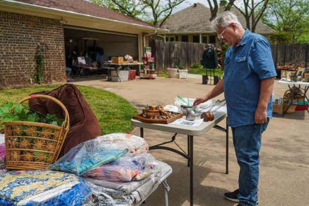 Man browsing items at a garage sale, standing near tables displaying household goods, clothing, and a basket. A garage filled with more items is visible in the background.