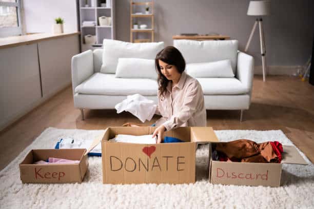 Woman sorts clothes into three labeled boxes: Keep, Donate, Discard, in a living room with a white couch and shelves in the background.