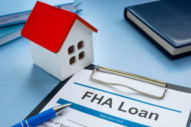 FHA loan application form on a clipboard next to a model house with a red roof and a blue pen on a desk.