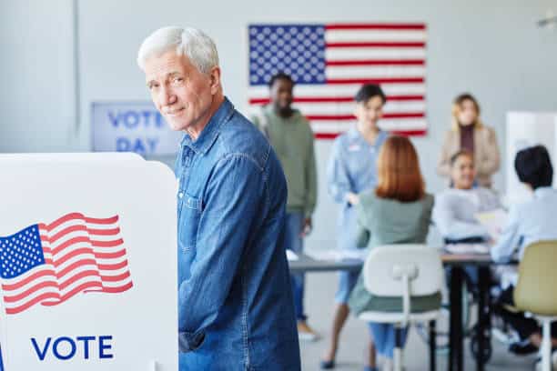 A man stands at a voting booth in front of a U.S. flag. Other people are seated and standing in the background in a polling station.