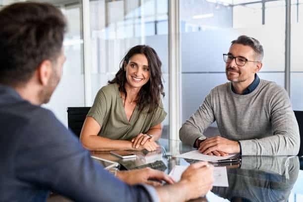 A woman and a man are sitting at a glass table, smiling and listening to another person who is speaking to them in a modern office setting.