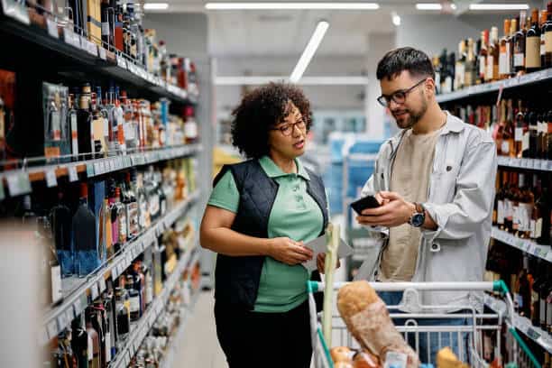 Two people in a store aisle are looking at a phone. A shopping cart with grocery items is in front of them. Shelves filled with bottles are in the background.