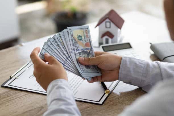 A person holding a fan of hundred-dollar bills at a desk with a contract, model house, and calculator.