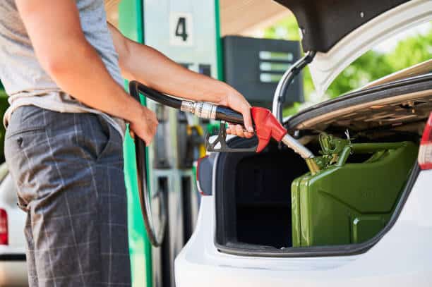 Person refueling a green gas canister placed in the open trunk of a car at a gas station.