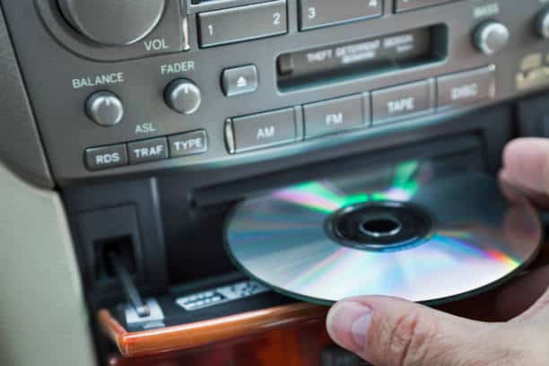 A hand loading a CD into a car stereo system featuring buttons for balance, fader, AM, FM, and tape.